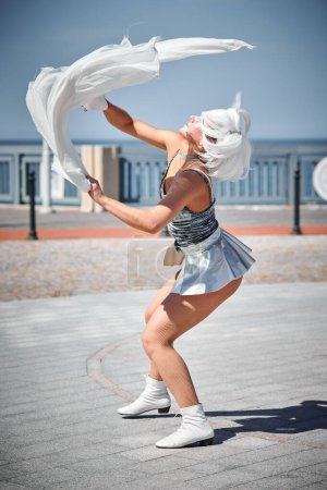 Young sexy girl in space silver micro skirt dancing with white silk scarf waving gracefully, female outdoor dance performance on seaside promenade creating an arousing outdoor spectacle