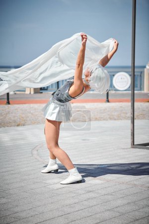 Foto de Joven chica sexy en el espacio micro falda de plata bailando con bufanda de seda blanca saludando con gracia, actuación de danza al aire libre femenina en el paseo marítimo creando un espectáculo al aire libre excitante - Imagen libre de derechos