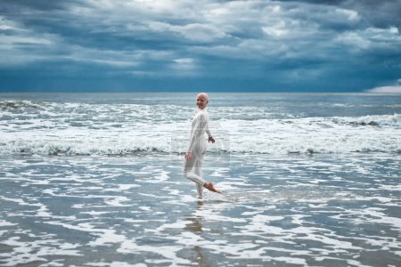 Happy hairless girl with alopecia in white futuristic suit dancing on beach bathed by ocean waves, performance of bald strong female artist, overcoming challenges of life and self confidence