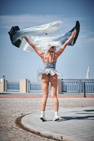 Foto de Joven chica sexy en el espacio micro falda de plata bailando con ventilador de rendimiento saludando con gracia, danza al aire libre femenina en el paseo marítimo creando espectáculo al aire libre armoniza con el ritmo de las olas del mar - Imagen libre de derechos