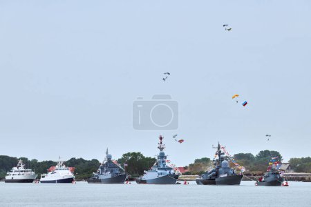 Photo for Airborne forces paratroopers flying Russian flag over naval forces parade warships along coastline, seafaring tradition of military ships formation at Navy Day, nautical spectacle of russian sea power - Royalty Free Image