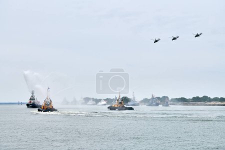 Photo for Fireboat sailing along Russian naval forces parade warships with military helicopters along coastline, seafaring tradition of military ships formation, nautical spectacle of russian sea power - Royalty Free Image