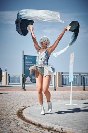 Foto de Joven chica sexy en el espacio micro falda de plata bailando con ventilador de rendimiento saludando con gracia, danza al aire libre femenina en el paseo marítimo creando espectáculo al aire libre armoniza con el ritmo de las olas del mar - Imagen libre de derechos