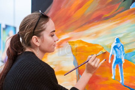 Atractiva pintora femenina apasionadamente dibuja cuadro con pincel para exposición callejera al aire libre, vista lateral de artista femenina infundiendo vida en el espacio de arte al aire libre