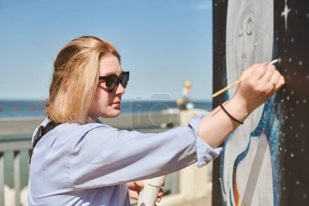 Junge Malerin mit Sonnenbrille malt leidenschaftlich Bild mit Pinsel für Straßenausstellung im Freien, Künstlerin in lebendige Kunstwerke bei strahlendem Sonnenschein vertieft