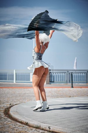 Junges sexy Mädchen in silbernem Mikrorock tanzt mit anmutig winkendem Ventilator, weiblicher Outdoor-Tanz auf der Strandpromenade schafft ein Outdoor-Spektakel, das mit dem Rhythmus der Meereswellen harmoniert