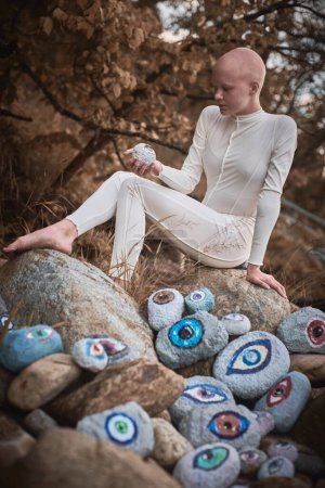 Jeune fille sans cheveux avec alopécie en costume futuriste blanc examine pensivement pierre avec oeil au paysage surréaliste, la performance symbolise l'introspection et la réévaluation de l'individualité