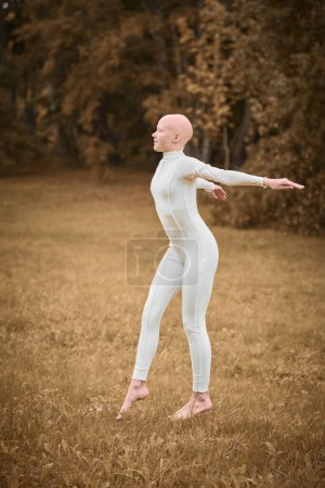 Full length portrait of young hairless girl ballerina with alopecia in tight white suit prepares to jump on fall lawn in park, symbolizing overcoming challenges and gracefully acceptance individuality