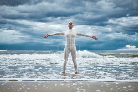 Photo for Happy hairless girl with alopecia in white futuristic suit stands with spread arms on beach bathed by ocean waves, performance of bald strong female artist symbolizing self acceptance - Royalty Free Image