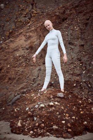 Full length portrait of young hairless girl with alopecia in white futuristic suit throws piece of barren soil thoughtfully, signifies vulnerability of planet and environmental issues