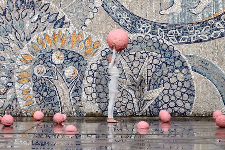 Young hairless girl with alopecia in white futuristic suit dancing outdoor smoothly holding pink ball on abstract mosaic Soviet background, symbolizes self expression and cultural identity