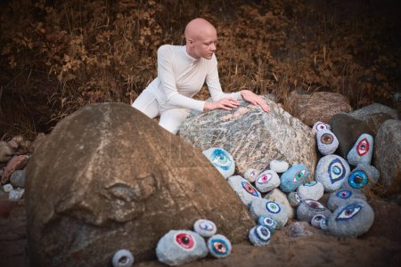 Junges haarloses Mädchen mit Alopezie im weißen futuristischen Kostüm blickt auf surreale Landschaft mit vielen Felsenaugen, die die Verbindung zwischen menschlichem Geist und Erde symbolisiert
