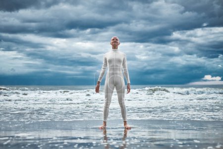 Photo for Happy hairless girl with alopecia in white futuristic suit stands on beach bathed by ocean waves, metaphoric performance of bald strong female artist, overcoming challenges of life and self confidence - Royalty Free Image