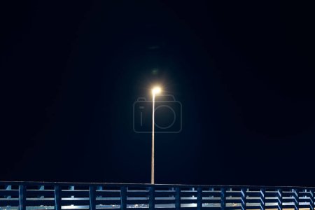 Einsamer Laternenpfahl wirft schummriges kaltblaues Glühen entlang der Ufermole, die vor pechschwarzer Leinwand des Nachthimmels steht, malt poetisches Bild der Mole in der Stille der Nacht, Gelassenheit einsamer Momente