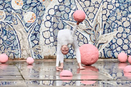 Tanz der jungen Ballerina mit Alopezie im weißen futuristischen Anzug mit plastischen und flexiblen Bewegungen zwischen rosa Kugeln auf abstraktem sowjetischen Mosaik, symbolisiert den Selbstausdruck