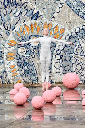 Junge haarlose Ballerina mit Alopezie im weißen futuristischen Anzug, die im Freien tanzt und zwischen rosa Kugeln auf abstraktem sowjetischen Mosaik springt, symbolisiert den Selbstausdruck