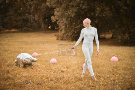 Retrato de niña sin pelo joven con alopecia en tela blanca caminando juguete tardígrado en el parque de otoño, escena surrealista con chica adolescente calva reflexionar sobre hilos entrelazados de la vida y el arte