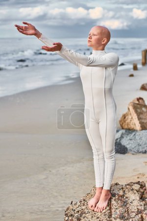 Ganzkörperporträt eines haarlosen Mädchens mit Alopezie im weißen futuristischen Anzug, das am steinernen Strand am Meer steht und die Arme ausstreckt. Metaphorische surreale Szene mit einem glatzköpfigen hübschen Teenager-Mädchen strahlt Hoffnung aus.