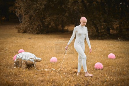 Porträt eines jungen haarlosen Mädchens mit Alopezie in weißem Tuch zu Fuß tardigrade Spielzeug im Herbst Park, surreale Szene mit glatzköpfigen Teenager-Mädchen reflektieren über verflochtene Fäden des Lebens und der Kunst