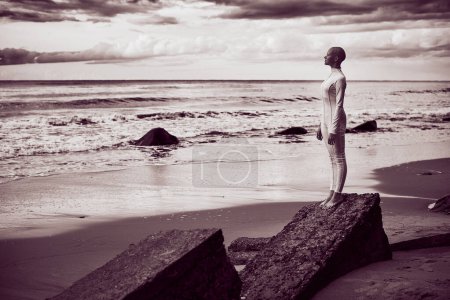 Ganzkörperporträt eines jungen haarlosen Mädchens mit Alopezie im weißen futuristischen Anzug, das am Strand am Meer steht, Schwarz-Weiß-Porträt, surreale Szene mit glatzköpfigem Teenager-Mädchen, dunkler Magenta-Ton