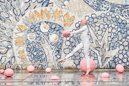Joven bailarina sin pelo con alopecia en traje futurista blanco bailando al aire libre y salta con esfera rosa sobre fondo abstracto mosaico soviético, simboliza la auto expresión