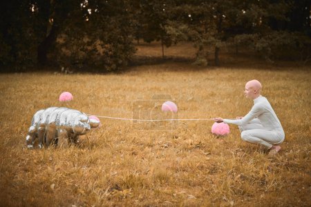Téléchargez les photos : Portrait de jeune fille sans cheveux avec alopécie en tissu blanc jouant avec un jouet tardif dans un parc d'automne, scène surréaliste avec une adolescente chauve réfléchir sur les fils entrelacés de la vie et de l'art - en image libre de droit