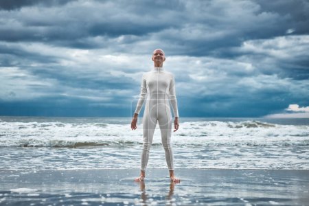 Happy hairless girl with alopecia in white futuristic suit stands on beach bathed by ocean waves, metaphoric performance of bald strong female artist, overcoming challenges of life and self confidence