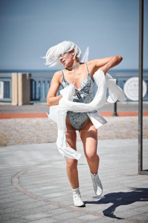 Young sexy girl in space silver micro skirt dancing with white silk scarf waving gracefully, female outdoor dance performance on seaside promenade creating an arousing outdoor spectacle