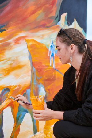 Atractiva pintora femenina apasionadamente dibuja cuadro con pincel para exposición callejera al aire libre, vista lateral de artista femenina infundiendo vida en el espacio de arte al aire libre