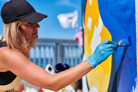 Erwachsene Malerin in schwarzer Mütze malt leidenschaftlich Bild mit Pinsel für Straßenausstellung im Freien mit lebendigen Farben, visuelles Spektakel durch ihre ausdrucksstarken Pinselstriche