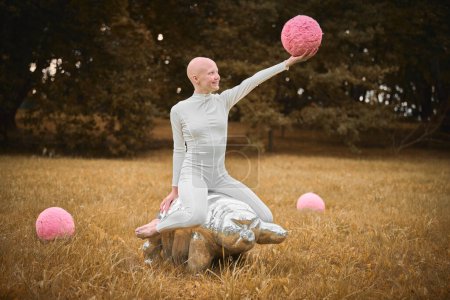 Joven chica sin pelo con alopecia en tela blanca se sienta en la figura tardía y sostiene la bola rosa en la mano en el parque de césped de otoño, escena surrealista con chica adolescente calva se involucra con elementos simbólicos