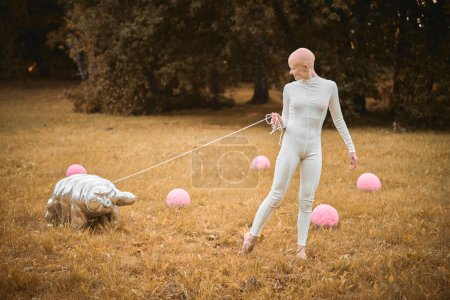 Photo for Portrait of young hairless girl with alopecia in white cloth walking tardigrade toy in fall park, surreal scene with bald teenage girl reflect on intertwining threads of life and art - Royalty Free Image