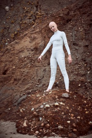 Ganztägiges Porträt eines jungen haarlosen Mädchens mit Alopezie im weißen futuristischen Anzug, das nachdenklich eine Handvoll karger Erde hält und damit das Potenzial für eine Koexistenz zwischen Mensch und Umwelt hervorhebt