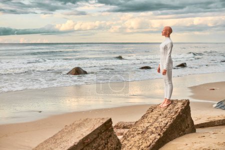 Ganztägiges Porträt eines jungen haarlosen Mädchens mit Alopezie im weißen futuristischen Anzug, das am steinernen Strand am Meer steht, metaphorische surreale Szene mit glatzköpfigem Teenager-Mädchen strahlt Zuversicht und einzigartige Schönheit aus