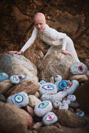 Joven chica sin pelo con alopecia en traje futurista blanco mirando paisaje surrealista con muchos ojos de rocas, simbolizando la conexión entre el espíritu humano y la tierra