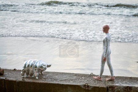 Young hairless girl with alopecia in white futuristic suit walking on concrete fence with toy tardigrade on sea background, bald pretty girl symbolizes courage and acceptance of unique appearance