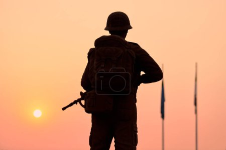 Téléchargez les photos : Vue arrière de la silhouette du soldat en équipement militaire tient l'arme contre le coucher de soleil sanglant et les drapeaux abaissés des pays, scène lourde symbolisant l'honneur solennel et les sacrifices faits au nom de la paix - en image libre de droit