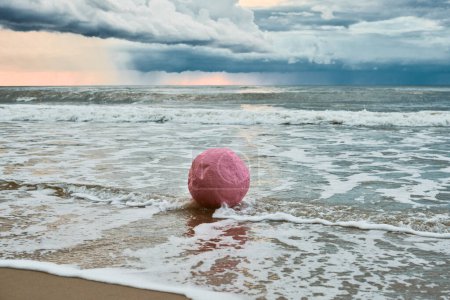 Große rosa Kugel umspült von Wellen gegen die Küste gegen Horizont und Gewitterwolken, rosa Ball Symbol der Gelassenheit Kontraste gegen brausende Turbulenzen am Himmel und unerbittliche Energie des Meeres
