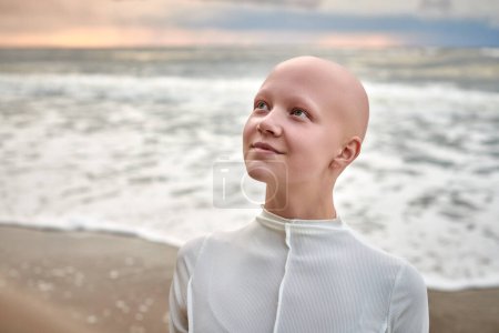 Photo for Close up portrait of smiling hairless girl with alopecia in white futuristic costume on sea background, bald pretty teenage girl showcasing unique beauty and identity with pride, unusual alien girl - Royalty Free Image