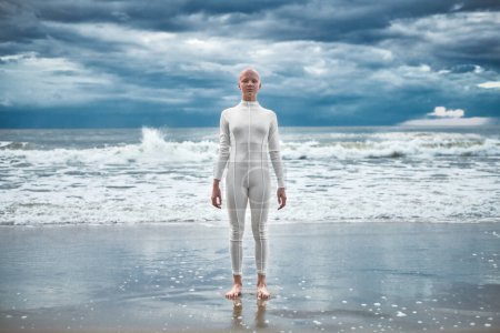 Happy hairless girl with alopecia in white futuristic suit stands on beach bathed by ocean waves, metaphoric performance of bald strong female artist, overcoming challenges of life and self confidence