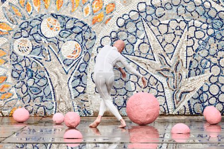 Junge haarlose Ballerina mit Alopezie im weißen futuristischen Anzug, die im Freien zwischen rosa Kugeln auf abstraktem sowjetischen Mosaik tanzt, symbolisiert Selbstausdruck und Akzeptanz einzigartiger Schönheit