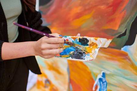 Pincel de mano de pintora femenina sumerge el pincel en una paleta de pinturas coloridas para la pintura en vivo de la imagen para la exposición callejera al aire libre, vista de cerca de la mano de artista femenina sosteniendo el pincel