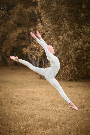 Photo for Full length portrait of young hairless girl ballerina with alopecia in tight white suit jumps on fall lawn in park, symbolizing overcoming challenges and gracefully acceptance individuality - Royalty Free Image
