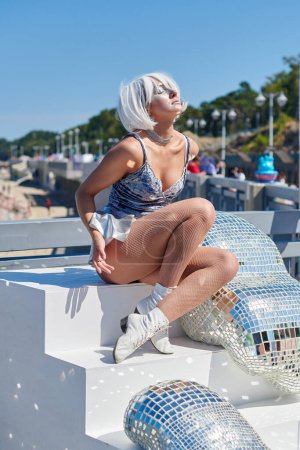 Foto de Joven chica sexy en el espacio micro falda de plata y peluca cósmica de plata se sienta en escaleras blancas junto a un objeto de arte espacial abstracto con un montón de pequeños espejos, simbolizando la ternura y la sexualidad femenina - Imagen libre de derechos