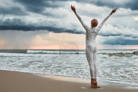 Haarloses Mädchen mit Alopezie im weißen futuristischen Anzug, das am Strand am Meer steht, ausgestreckte Arme zum wolkenverhangenen Himmel, Ganzkörperporträt von der Rückseite, metaphorische Performance mit glatzköpfigem sensiblen Teenager-Mädchen