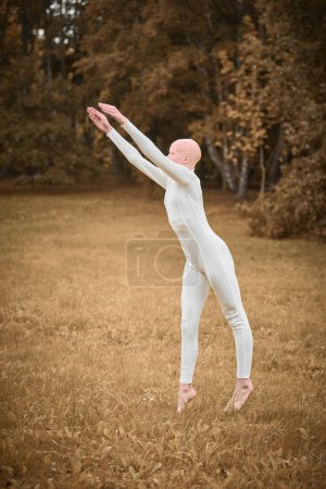 Retrato de larga duración de la bailarina joven sin pelo con alopecia en traje blanco apretado se prepara para saltar en el césped de otoño en el parque, lo que simboliza la superación de los desafíos y la individualidad aceptación con gracia