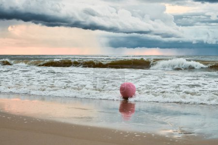 Large pink sphere lapped by waves against seashore against horizon and storm clouds, pink ball symbol of serenity contrasts against brewing turbulence in sky and relentless energy of sea