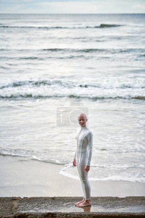 Junges haarloses Mädchen mit Alopezie im weißen futuristischen Anzug steht auf Betonzaun auf Meeresgrund, Glatze hübsches Teenager-Mädchen symbolisiert Mut und Akzeptanz der einzigartigen Erscheinung