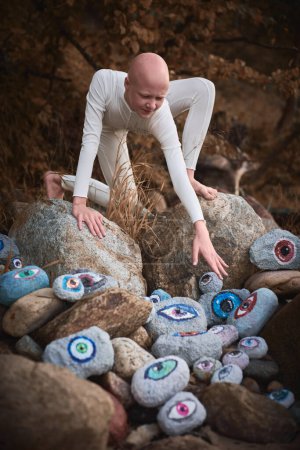 Young hairless girl with alopecia in white futuristic costume reaches hand for surreal landscape with lot of rocks with eyes, profound symbolism of embracing individuality in surreal world