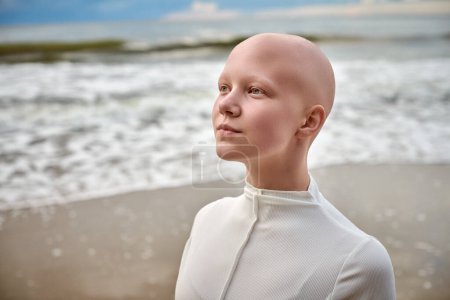 Close up portrait of young hairless girl with alopecia in white futuristic costume on sea background, bald pretty teenage girl showcasing unique beauty and identity with pride, unusual alien girl